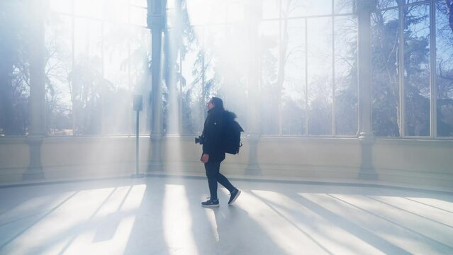 Young Latin woman tourist in warm clothes with professional photo camera walking through the mist and the rays of light and standing inside of The Glass Palace in Madrid, Spain 