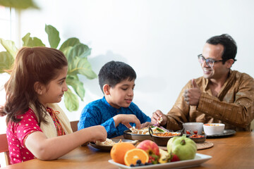 Happy Indian father with daughter and son having meal at home, family in traditional dress tasting...