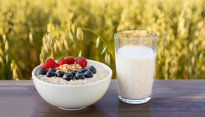 oatmeal with berries in bowl and glass of vegan oat milk on table over against ripe cereal field on the background. Healthy breakfast concept