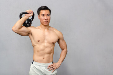 Asian strong handsome young male muscular shirtless fitness model in sporty shorts standing smiling holding lifting metal kettlebell dumbbell showing biceps triceps muscle in gym on gray background
