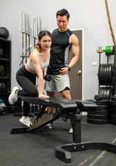 Asian strong young shirtless male muscular fitness trainer in sporty shorts standing helping saving female athlete in sexy sport bra lifting up metal dumbbells sitting on bench working out in gym