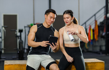 Asian strong young fit male muscular fitness athlete model and female friend sitting take break drink water wipe sweat on wood box smiling talking together showing schedule on tablet computer in gym