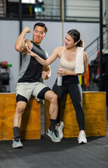 Fototapeta na wymiar Asian strong young fit male muscular fitness athlete model showing off bicep muscle to female friend sitting take break drink water wipe sweat on wood box smiling hold thumb up complimenting in gym