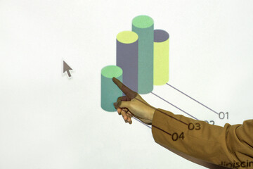 The confident stance and precise pointing suggest an educational or business-oriented discussion unfolding in an office or conference room setting. Hand pointing at graph on presentation monitor.