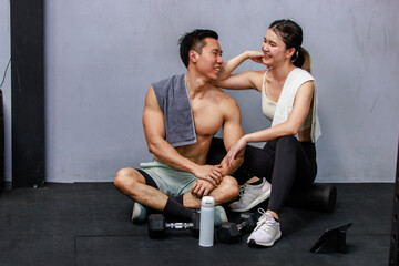 Obraz premium Asian strong young male muscular shirtless fitness model in sporty shorts and female athlete in sport bra sitting smiling taking break holding water bottle talking together in gym on gray background
