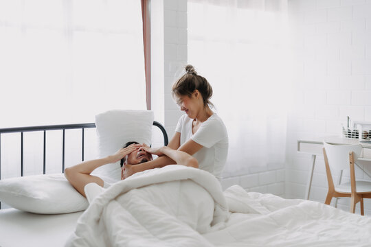 Funny asian couple wife try to wake lazy husband up in white bedroom.