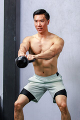 Asian strong handsome young male muscular shirtless fitness model in sporty shorts standing smiling holding lifting metal kettlebell dumbbell showing biceps triceps muscle in gym on gray background