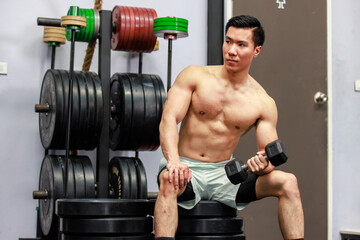 Fototapeta na wymiar Millennial Asian fit strong handsome shirtless muscular male fitness model athlete in shorts sitting on barbell weights lifting training exercising alone in gym.