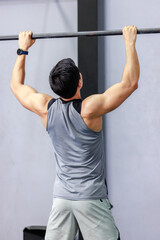 Asian strong unrecognizable unknown young shirtless male muscular fitness model in sporty shorts holding pulling up gymnastic rings lifting working out back muscle strength in gym on gray background