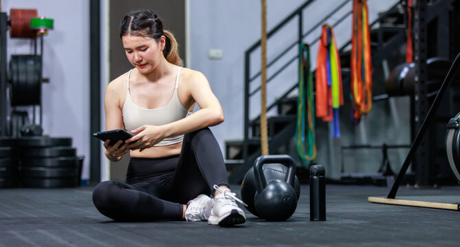 Millennial Asian strong young fit female muscular fitness athlete model in sportswear sport bra and legging sitting on floor taking break from working out checking message on touchscreen smartphone - Powered by Adobe