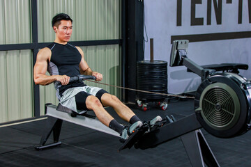 Fototapeta na wymiar Asian strong young fit male muscular fitness athlete model in sleeveless shirt and sporty shorts sitting using heavy cable pulling machine working out training exercising in gym.