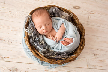A cute newborn boy in the first days of life sleeps in a blue winding. Wicker brown basket on a light wood background.