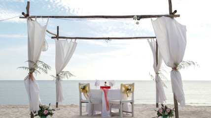 Romantic dinner table set and chair preparation with white theme decoration, white sand, clear cloudy sky, and tranquil, peaceful, beautiful beach view at the seashore. Good place for wedding proposal