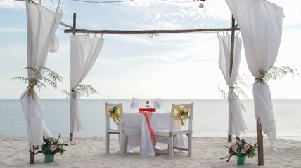 Romantic dinner table set and chair preparation with white theme decoration, white sand, clear cloudy sky, and tranquil, peaceful, beautiful beach view at the seashore. Good place for wedding proposal