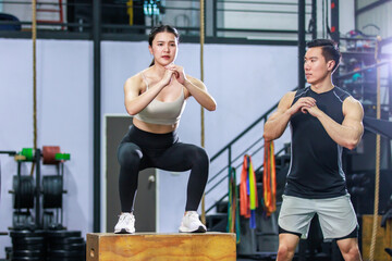 Naklejka premium Asian strong young fit male trainer helping teaching female muscular fitness athlete model in sexy sport bra and legging training exercising squatting on wooden box in gym full of exercise equipment