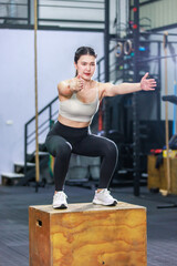 Millennial Asian fit strong muscular female fitness model athlete in sport bra legging and sneakers working out training exercising squatting on wooden box alone in gym full of exercise equipment