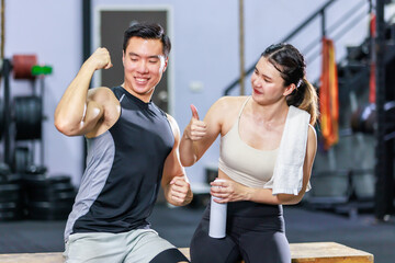 Fototapeta na wymiar Asian strong young fit male muscular fitness athlete model showing off bicep muscle to female friend sitting take break drink water wipe sweat on wood box smiling hold thumb up complimenting in gym