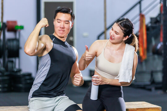 Asian strong young fit male muscular fitness athlete model showing off bicep muscle to female friend sitting take break drink water wipe sweat on wood box smiling hold thumb up complimenting in gym