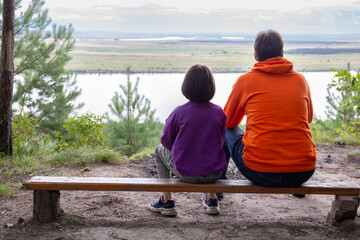 Dad and daughter sit on a bench with their backs to the camera and admire the beautiful natural...