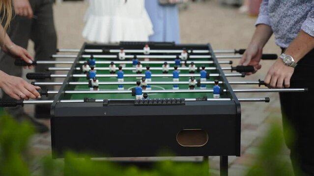 Man and woman play kicker against each other outdoors during disco at party. Table football for entertainment of guests at events, people are dancing at party nearby, players turn knobs with players.