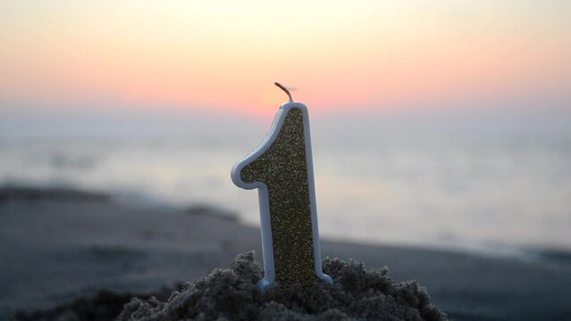 Numeral one from paraffin wax with yellow glitters burns on sandy beach of sea shore at dawn sunset close-up. Paraffin candle burning near blue sea. Concept holiday Birthday New Year party celebrating