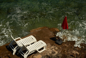 Titolo: On the tip of a rocky cliff, two comfortable white deckchairs and a red umbrella, are we ready for a swim?