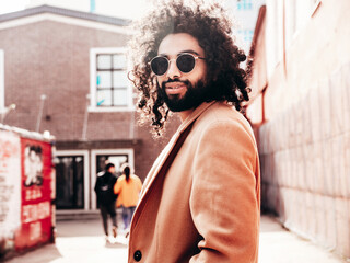 Naklejka na ściany i meble Handsome hipster model. Unshaven Arabian man dressed in brown coat clothes. Fashion male with long curly hairstyle posing outdoors in street. Cheerful and happy. At sunny day. In sunglasses