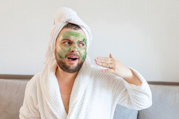 Surprised guy spends hours treating and caring for his skin, applies green face mask to pamper himself, wearing a soft bathrobe and a towel on his head - Powered by Adobe
