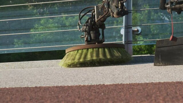 Green rotating city cleaner brushes sweeps away dirt from bridge. Municipal mud sweeper machine cleaning asphalt on bridge near city park