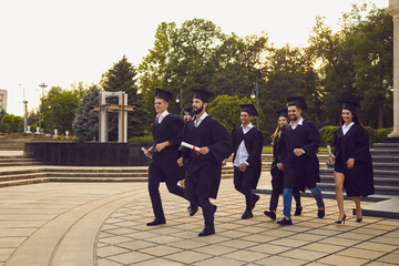 Successful graduation from university. Group of cheerful university graduates walking with dimplomas in hands in park together and celebrating graduation
