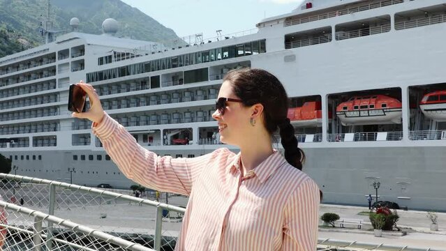 Portrait of young smiling woman making video call with smartphone. Huge cruise liner in background