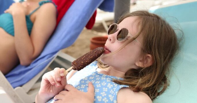 Teen girl in sunglasses eats ice cream lying on sun lounger on seaside near mother. Resting schoolgirl shrugs after tasting dessert. Careless holiday