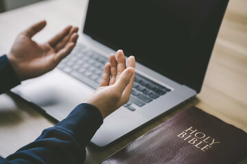 Christian hands folded for prayer with a holy bible and laptop on a wooden table in church for...