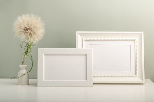 Two white empty photo frames and a small white vase with a dandelion on the table. Minimalistic interior.