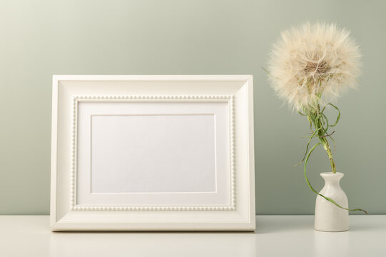White blank photo frame and small white vase with dandelion on the table. Minimalistic interior.