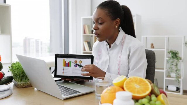Focused multicultural expert with meal plan on tablet conducting web meeting using portable laptop at work. Skilful female nutritionist explaining details of slimming program for client online.