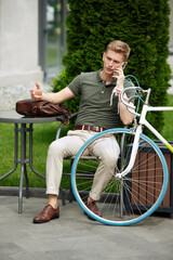 Obraz premium Stylish, handsome, serious businessman sitting on bench and talking on mobile phone. Going to work on bike. Solving business tasks. Concept of business, active lifestyle, fashion, youth, ecology