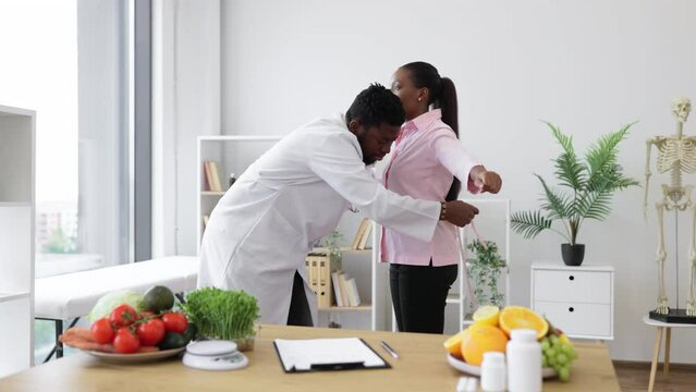Multiethnic man in doctor's coat finding out waist length of female patient with measuring tape in consulting room. Efficient dietitian calculating body mass index for assessing healthy weight.