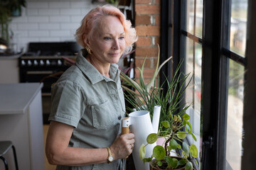 Senior woman at home watering indoor plants and looking out of window thinking