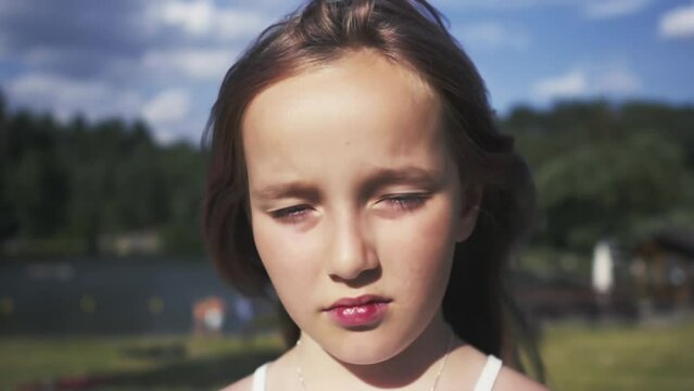 Portrait of an eight-year-old girl squinting her eyes on a bright summer day. In the background a grassy beach by a lake.