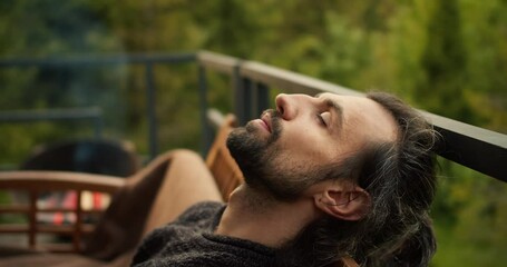 Close-up shot: A young brunette man with closed eyes is resting on a sofa on the balcony of a country house overlooking the mountains and coniferous forest. Spiritual rest, outdoor leisure - Powered by Adobe