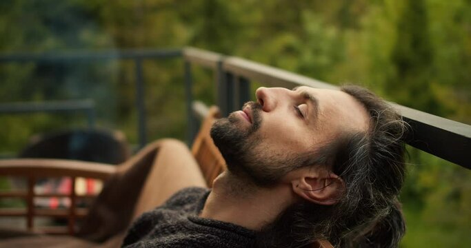 Close-up shot: A young brunette man with closed eyes is resting on a sofa on the balcony of a country house overlooking the mountains and coniferous forest. Spiritual rest, outdoor leisure