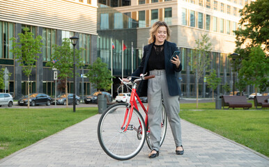 Business woman with smartphone on a bicycle in the city park near the office building