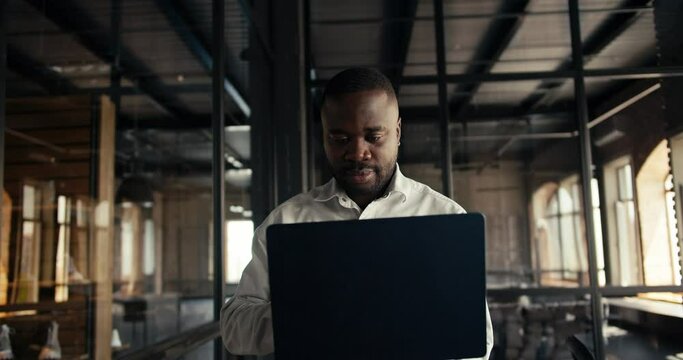 A man with Black skin in a white shirt with a laptop in his hands walks around the office in a loft style and works
