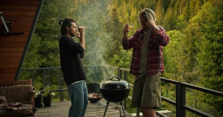 Two guys dance and have fun while cooking on the grill on a spacious balcony overlooking coniferous ice and mountains. Picnic fun, happy leisure