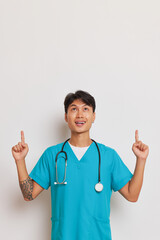 Asian male medical worker in blue uniform looks up and points with index fingers up posing on white background, professional people concept, copy space