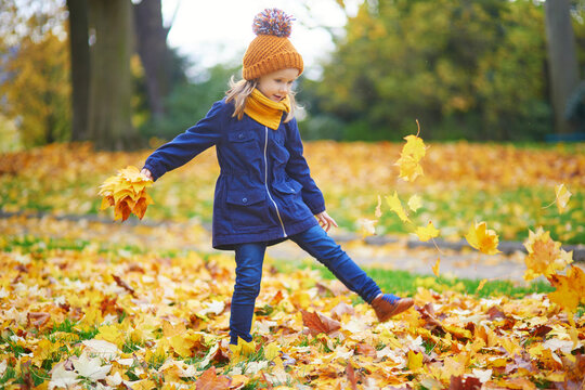 Adorable preschooler girl walking and kicking fallen leaves in autumn park in Paris, France