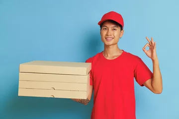Foto op Plexiglas Joyful guy dressed in red cap and red t-shirt posing on blue background with pizza boxes in one hand and shows ok sign with other hand, tasty food concept, copy space © South House Studio