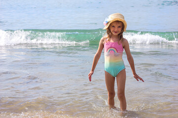 A beautiful little girl in a bright swimsuit and a hat stands against the backdrop of the sea on a...