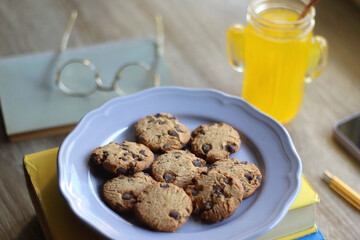 Plate of chocolate chip cookies, stack of books, reading glasses, orange soda, mobile phone and pen on the table. Hygge at home. Pastel colors, selective focus.